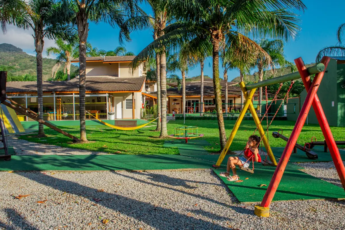 Menina brincando no balanço do parquinho, com gramado, coqueiros e o prédio do hotel ao fundo